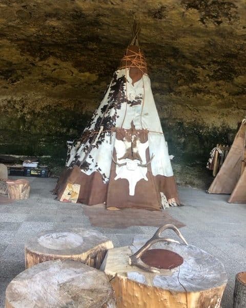 Tente tipie traditionnelle sous une grotte, entourée de troncs en bois.