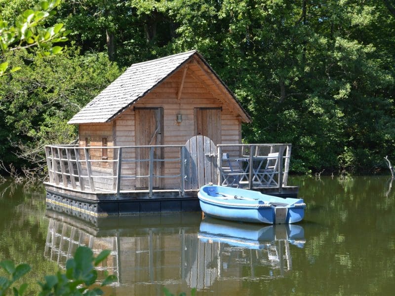 Cabane sur leau avec terrasse, entourée de verdure et bateau à rames.