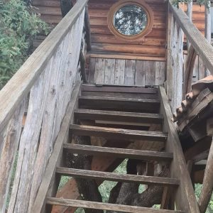 Cabane perchée en Corse, accès par un escalier en bois et fenêtre ronde.