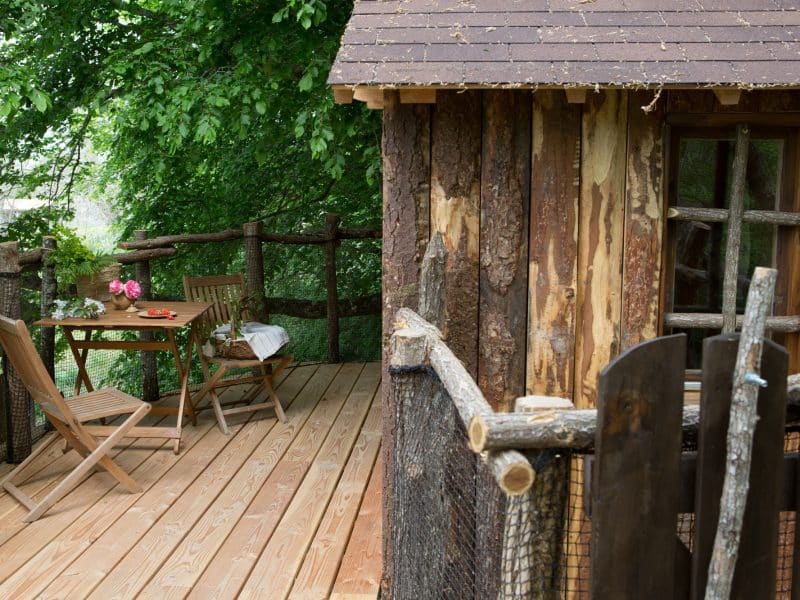 Cabane en bois perchée, avec terrasse en bois et table pour deux sous les arbres.
