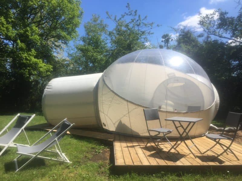 Hébergement insolite : bulle transparente sur une terrasse en bois, entourée de verdure.