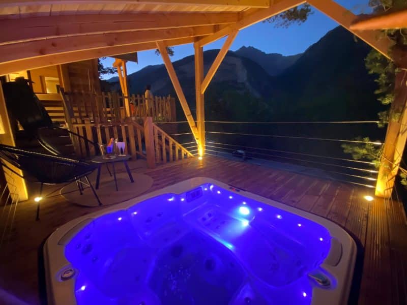 Chalet en bois avec jacuzzi lumineux, vue imprenable sur les montagnes au crépuscule.