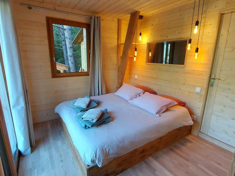 Chalet en bois avec lit douillet et grandes fenêtres lumineuses.