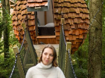 Cabane bulle en bois suspendue, entourée darbres, accès par un pont.