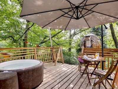 Cabane dans les arbres avec jacuzzi, terrasse en bois entourée de verdure.