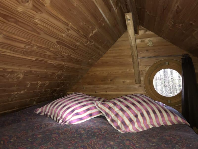 Cabane en bois avec un lit douillet et une fenêtre ronde offrant une vue sur la nature.