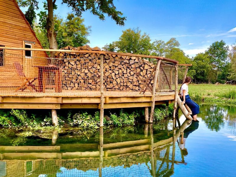 Cabane sur pilotis en bois, au bord dun étang, entourée de verdure.
