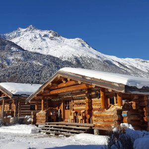Chalet en bois, entouré de neige, avec montagnes majestueuses en arrière-plan.
