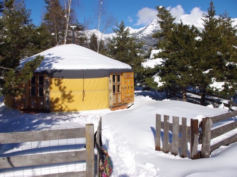 Yourte chaleureuse entourée de neige, avec une porte en bois sculpté et des arbres verdoyants.