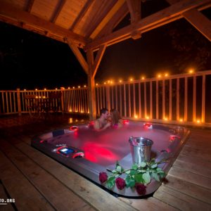 Cabane en bois avec jacuzzi romantique, éclairage doux et roses sur la terrasse.