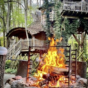 Cabane perchée en forêt, avec un feu de camp crépitant devant.