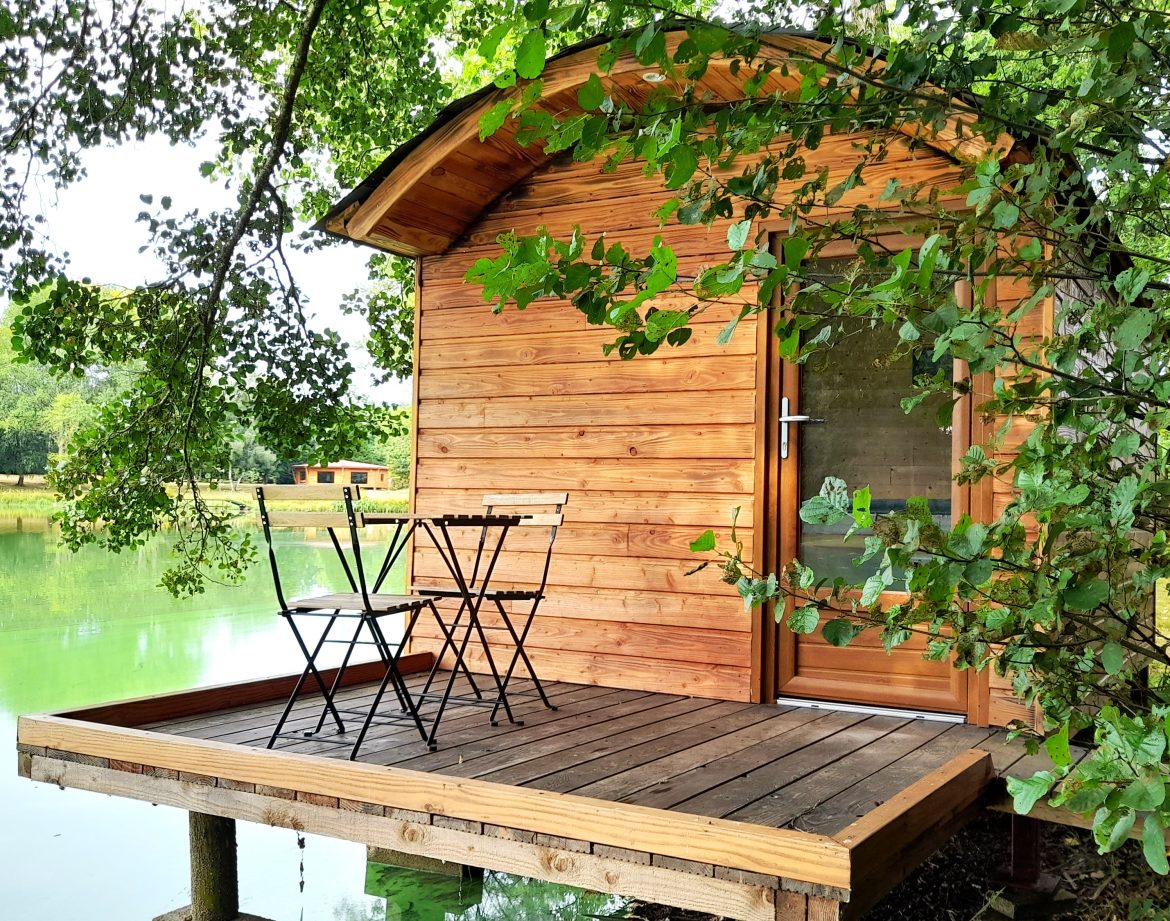 Cabane en bois sur pilotis au bord dun lac, avec une terrasse et deux chaises.