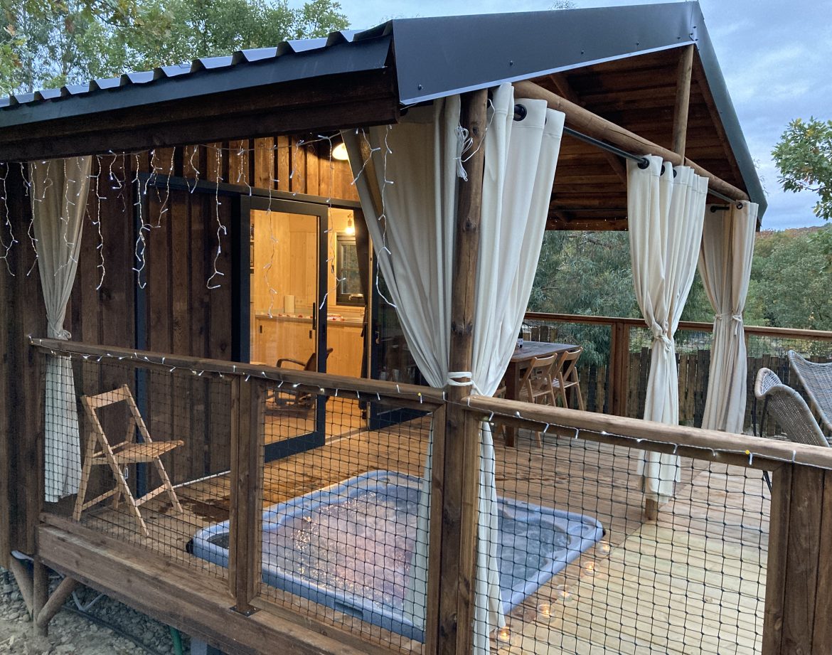 Cabane en bois avec jacuzzi, entourée de verdure et décorée de guirlandes lumineuses.