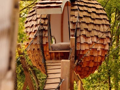 Cabane en bois en forme de sphère, perchée dans les arbres, accessible par un pont suspendu.