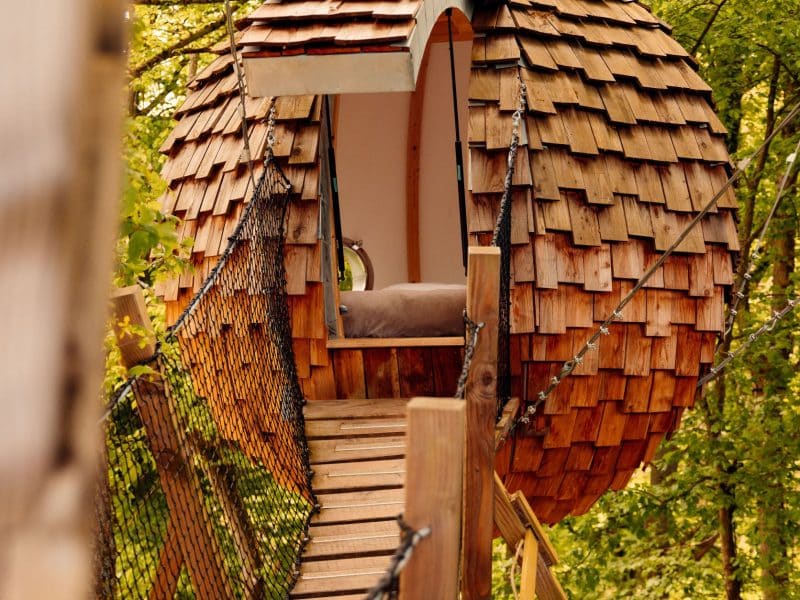 Cabane en bois en forme de sphère, perchée dans les arbres, accessible par un pont suspendu.