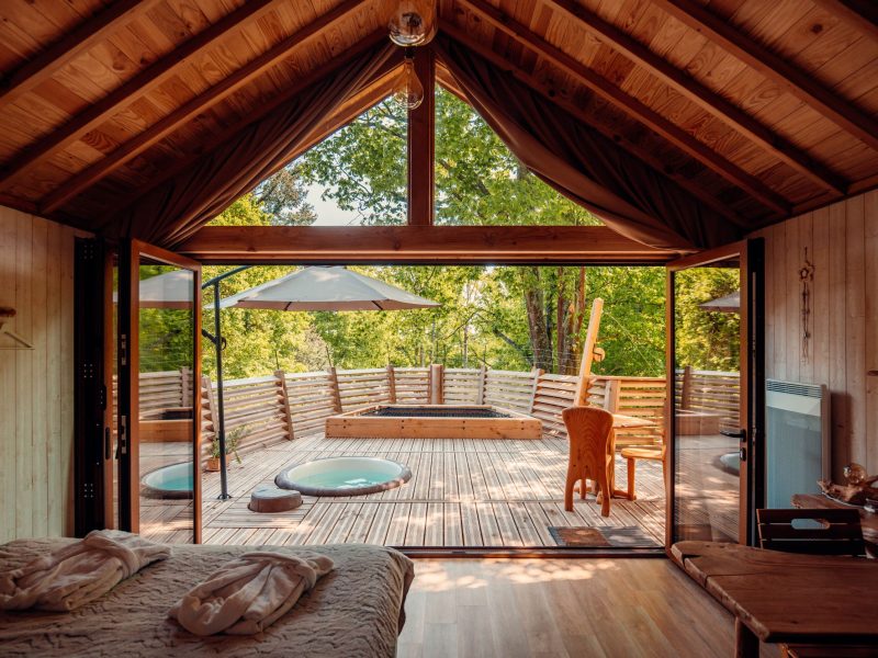 Cabane en bois avec terrasse ensoleillée et jacuzzi, vue sur la nature.