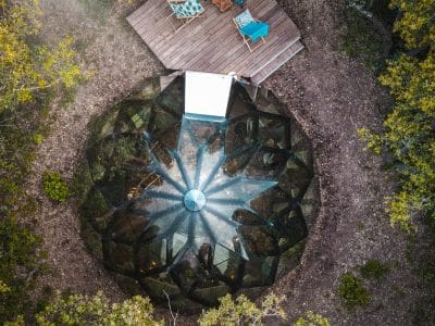 Hébergement insolite en verre, entouré darbres, avec terrasse en bois et chaises confortables.