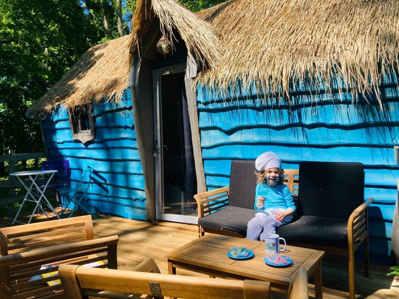 Cabane colorée avec toit de chaume, terrasse accueillante et enfant souriant.