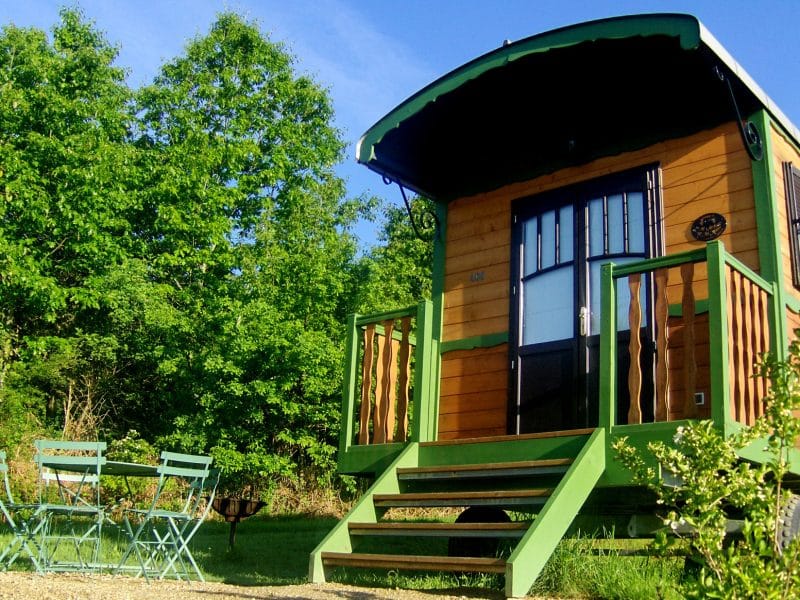 Charmante roulotte en bois avec terrasse et chaises vertes, entourée de verdure.