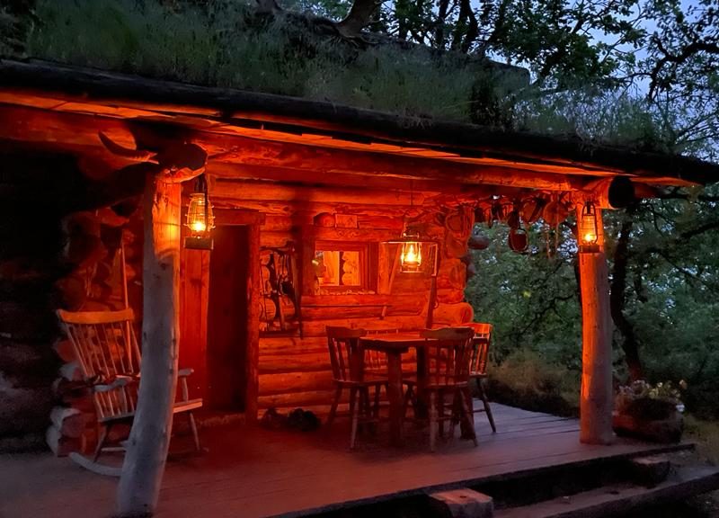 Cabane en bois éclairée par des lanternes, entourée de verdure au crépuscule.