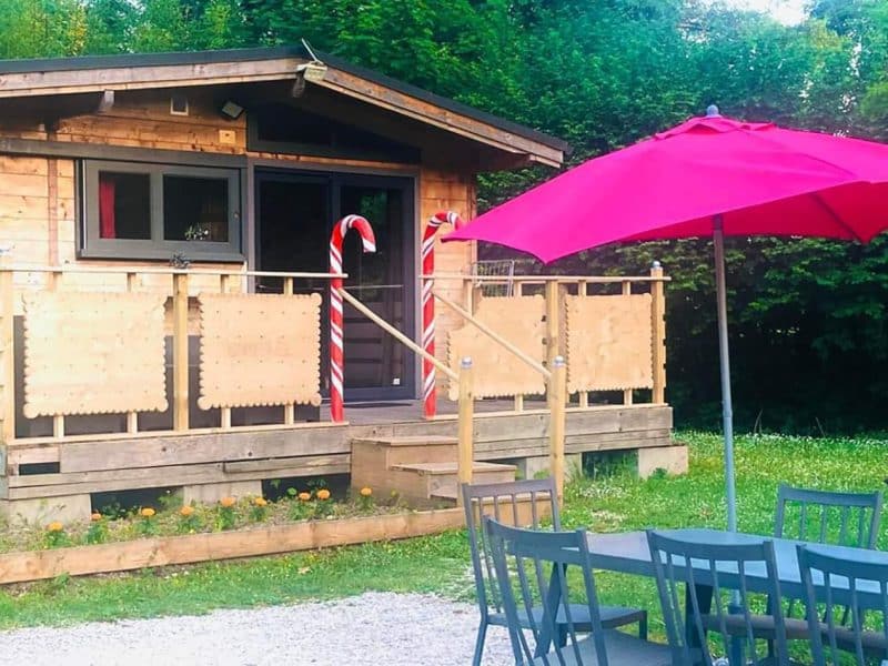 Chalet en bois avec terrasse, parasol rose et barbecue dans un cadre verdoyant.