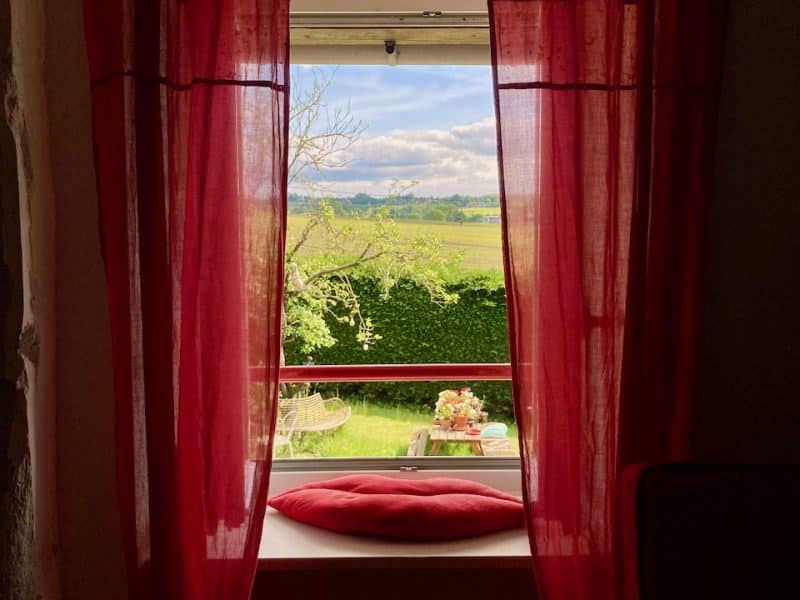 Chambre dhôtes avec rideaux rouges, vue sur un paysage verdoyant et ensoleillé.
