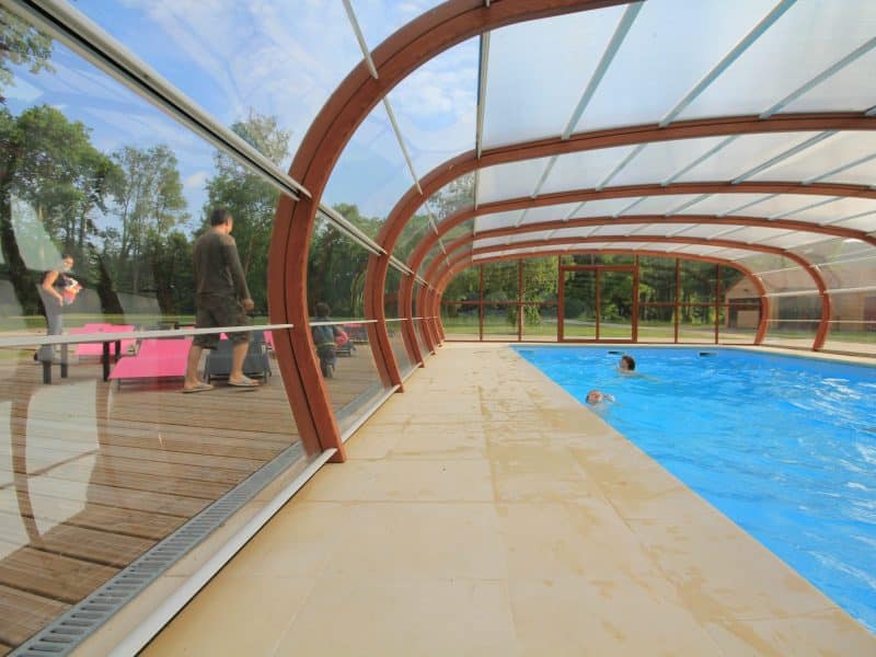 Gîte moderne avec piscine intérieure, entouré de verdure et de lumière naturelle.