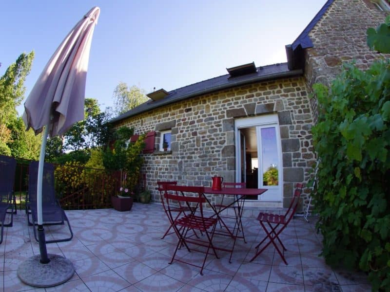 Gîte en pierre avec terrasse, chaises rouges et parasol, entouré de verdure.