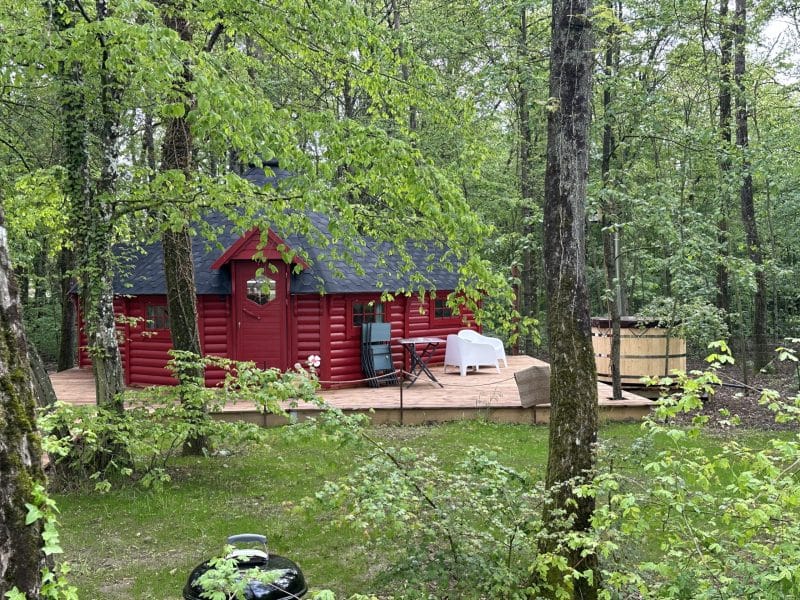 Cabane en bois rouge entourée darbres, avec terrasse et mobilier extérieur.
