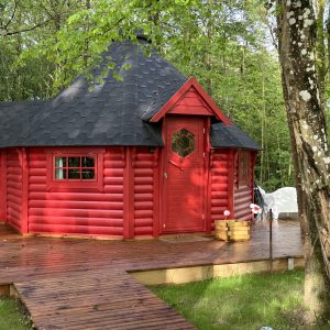 Cabane rouge en bois, entourée de verdure, avec un toit en ardoise et une porte hexagonale.