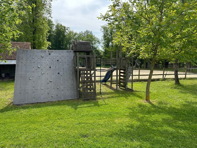 Aire de jeux en bois pour enfants, avec mur descalade et toboggan, entourée darbres.