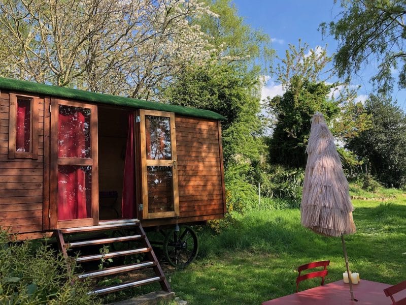 Roulotte en bois à Basse-Normandie, entourée de verdure et dun parasol.