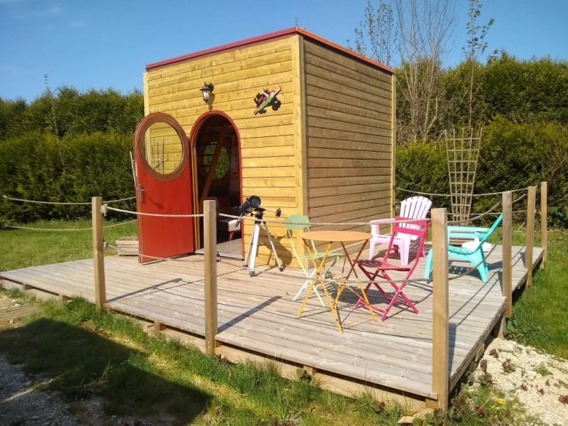 Cabane en bois avec terrasse en bois et chaises colorées, entourée de verdure.