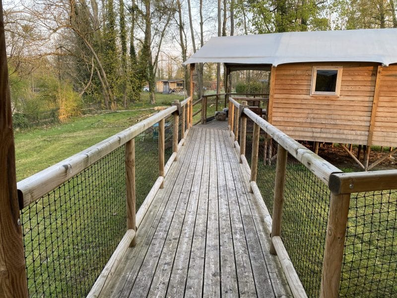 Cabane en bois sur pilotis, accessible par un chemin en bois entouré de verdure.