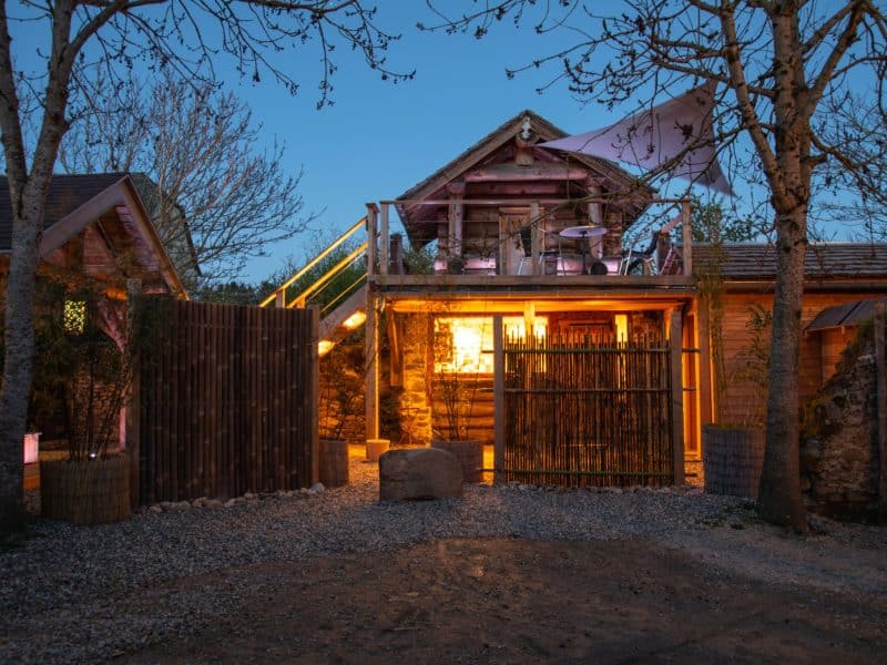 Cabane en bois illuminée, entourée darbres, offrant un cadre unique en Midi-Pyrénées.