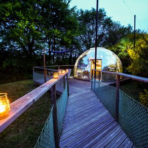 Dôme transparent en Bretagne, accessible par un chemin en bois, éclairé par des lanternes.