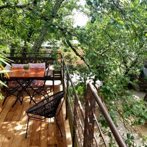 Cabane perchée dans les arbres, avec terrasse en bois et chaises noires.