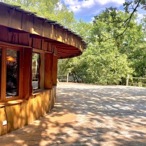 Cabane en bois ronde, entourée de verdure, avec une grande terrasse ensoleillée.