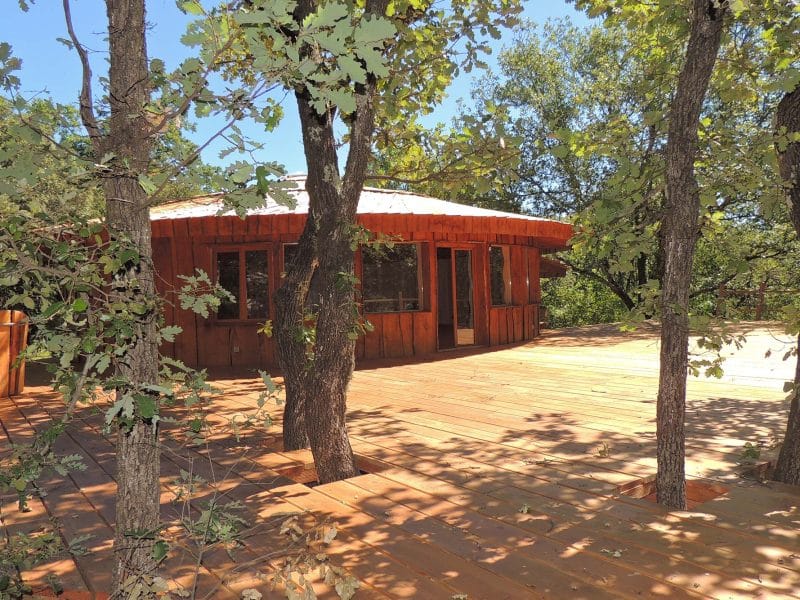 Cabane en bois au milieu des arbres, avec une grande terrasse ensoleillée.