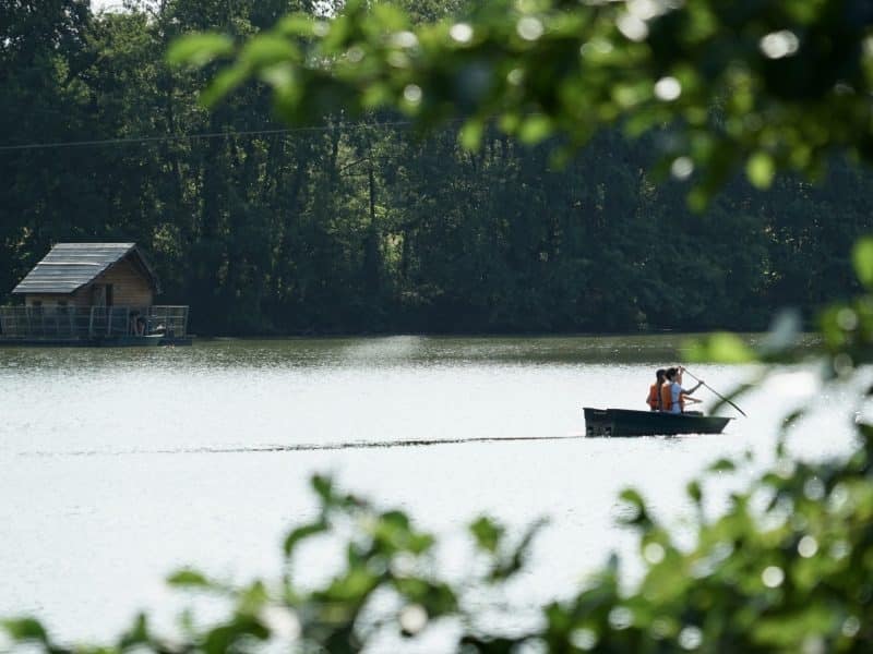 Cabane au bord de leau, avec un couple en barque sur un lac paisible.