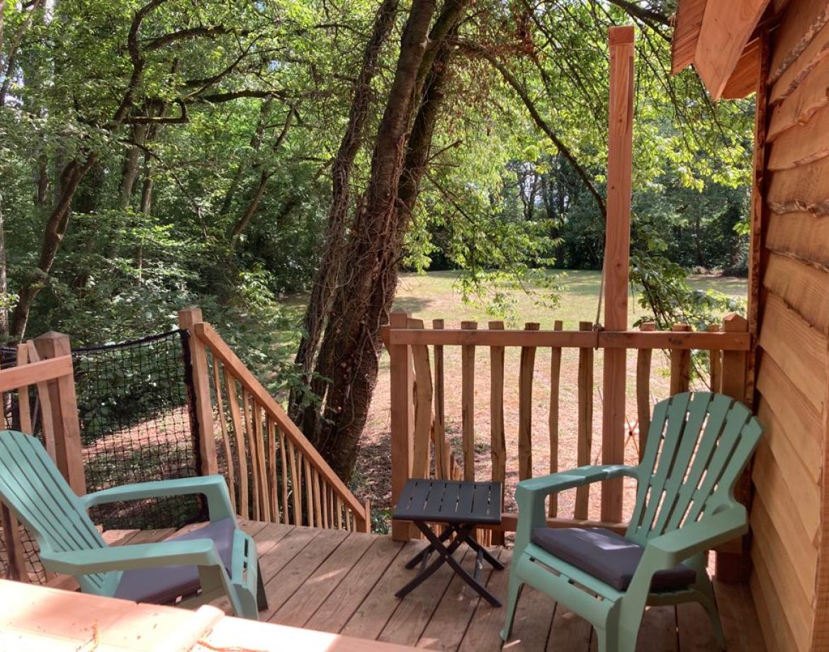 Cabane en bois dans les arbres, avec terrasse et chaises colorées, entourée de verdure.