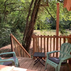 Cabane en bois dans les arbres, avec terrasse et chaises colorées, entourée de verdure.