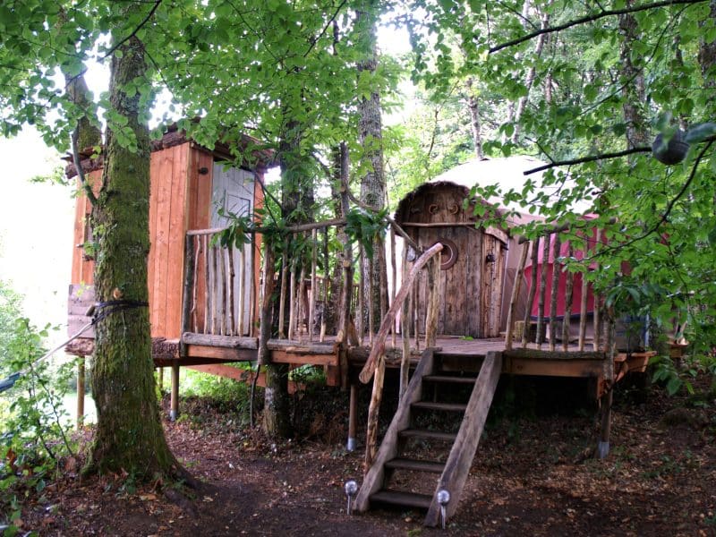 Cabane perchée dans les arbres, entourée de verdure, avec une terrasse en bois.