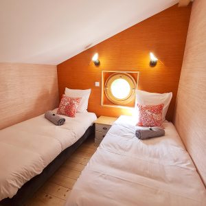 Chambre dhôtes atypique avec lits jumeaux et fenêtre ronde, ambiance chaleureuse.