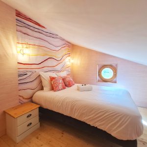 Chambre cosy dans un hébergement insolite, avec un mur coloré et une fenêtre ronde.