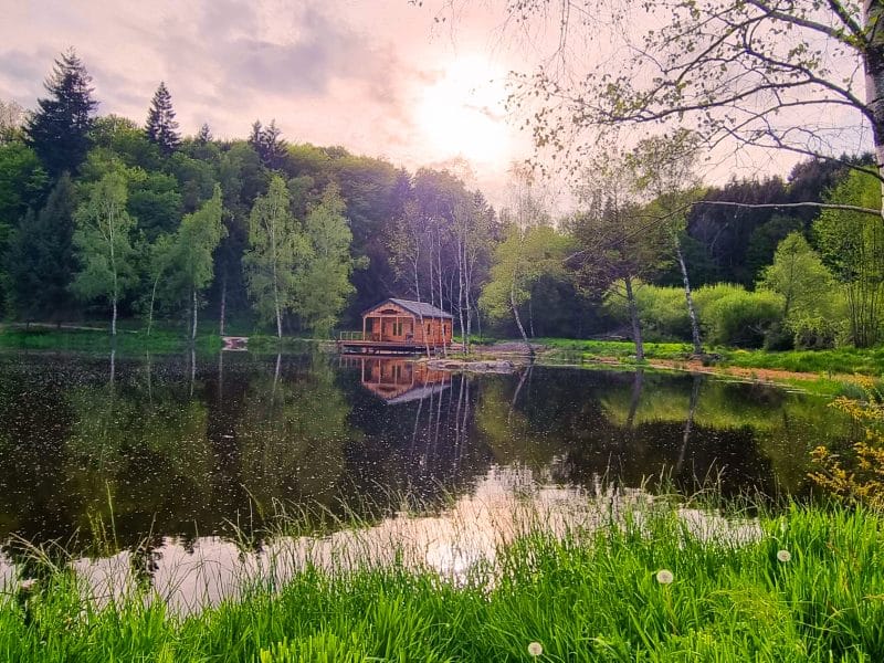 Cabane au bord dun lac, entourée de verdure et reflétant la lumière du soleil.