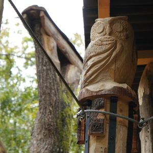 Cabane perchée en bois avec une sculpture de hibou, entourée darbres verdoyants.