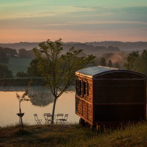 Roulotte au bord dun lac, entourée de verdure, offrant une vue paisible au lever du soleil.