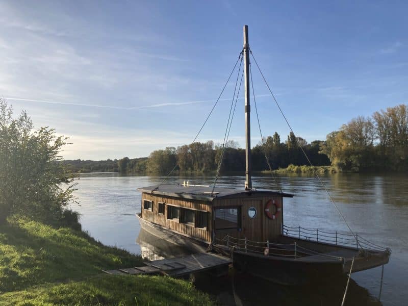 Maison flottante en bois au bord dune rivière, entourée de verdure.