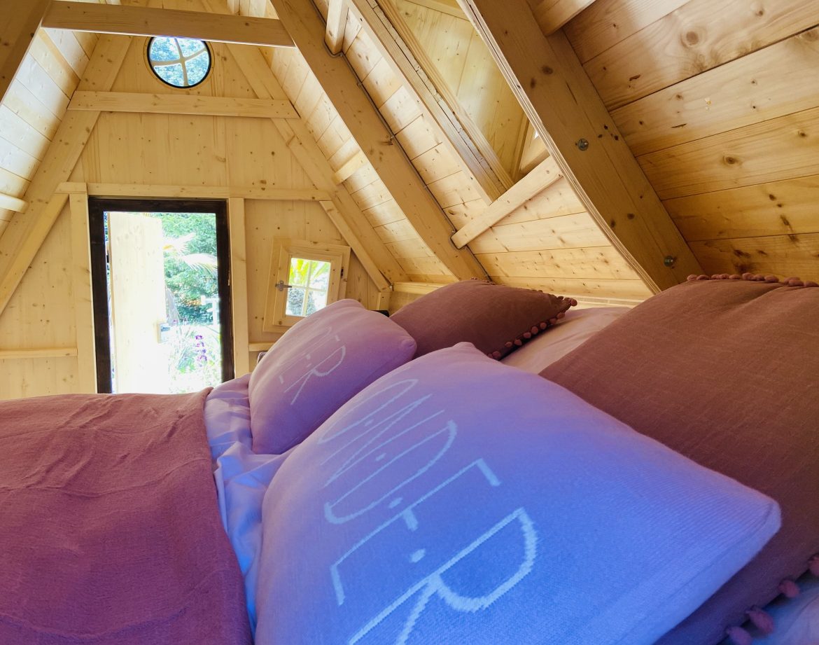 Cabane en bois en Bretagne, lit douillet avec coussins colorés et vue sur la nature.